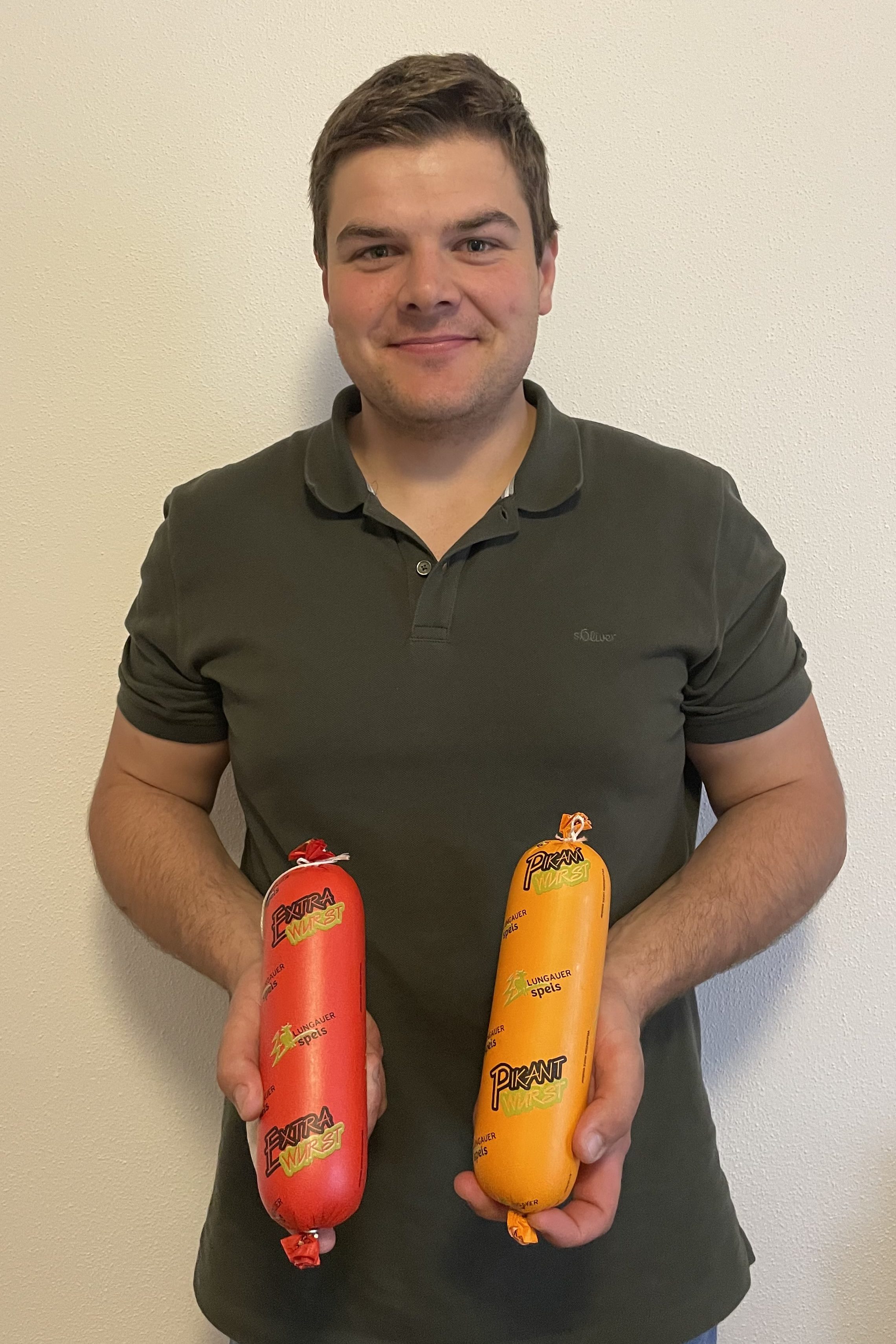 Mann hält zwei regionale Wurstwaren aus dem Lungau vor weißer Wand